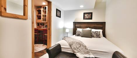 A bedroom with a large bed, a wooden headboard, a bench, a mirror, and a door leading to a dining area.
