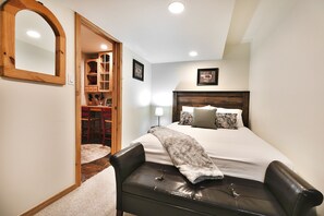 A bedroom with a large bed, a wooden headboard, a bench, a mirror, and a door leading to a dining area.