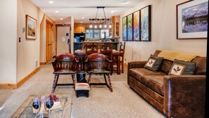 A spacious living room with a brown leather sofa, wooden dining table, and chairs. A kitchen is visible in the background with wooden cabinets and a large window.
