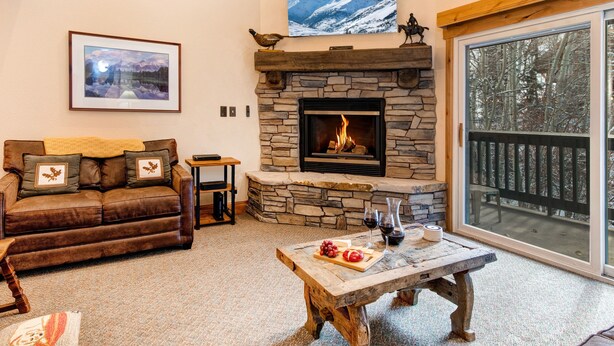 A cozy living room with a stone fireplace, a large window with a view of trees, a brown leather sofa, and a rustic coffee table.