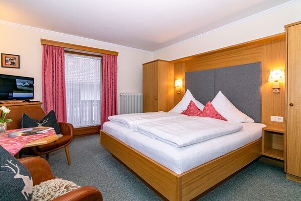 A hotel room with a wooden bed, a desk with a chair, a television, and a window with curtains. - Ferienhaus Ostara Balk/terr., 6 Schlafz.m. Du/wc, 1 Wohnz., Aufenthaltsraum, Max. 12 Pers (Ruhpolding)