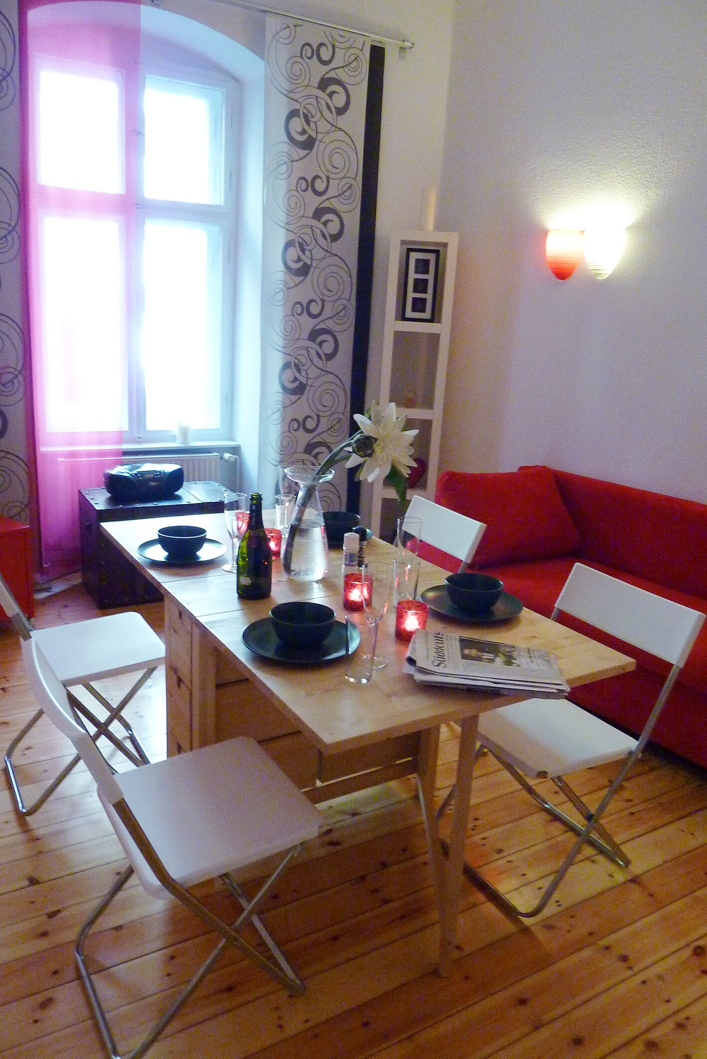 Un comedor con una mesa de madera dispuesta para dos personas, sillas blancas, un sofá rojo y un jarrón con una flor.