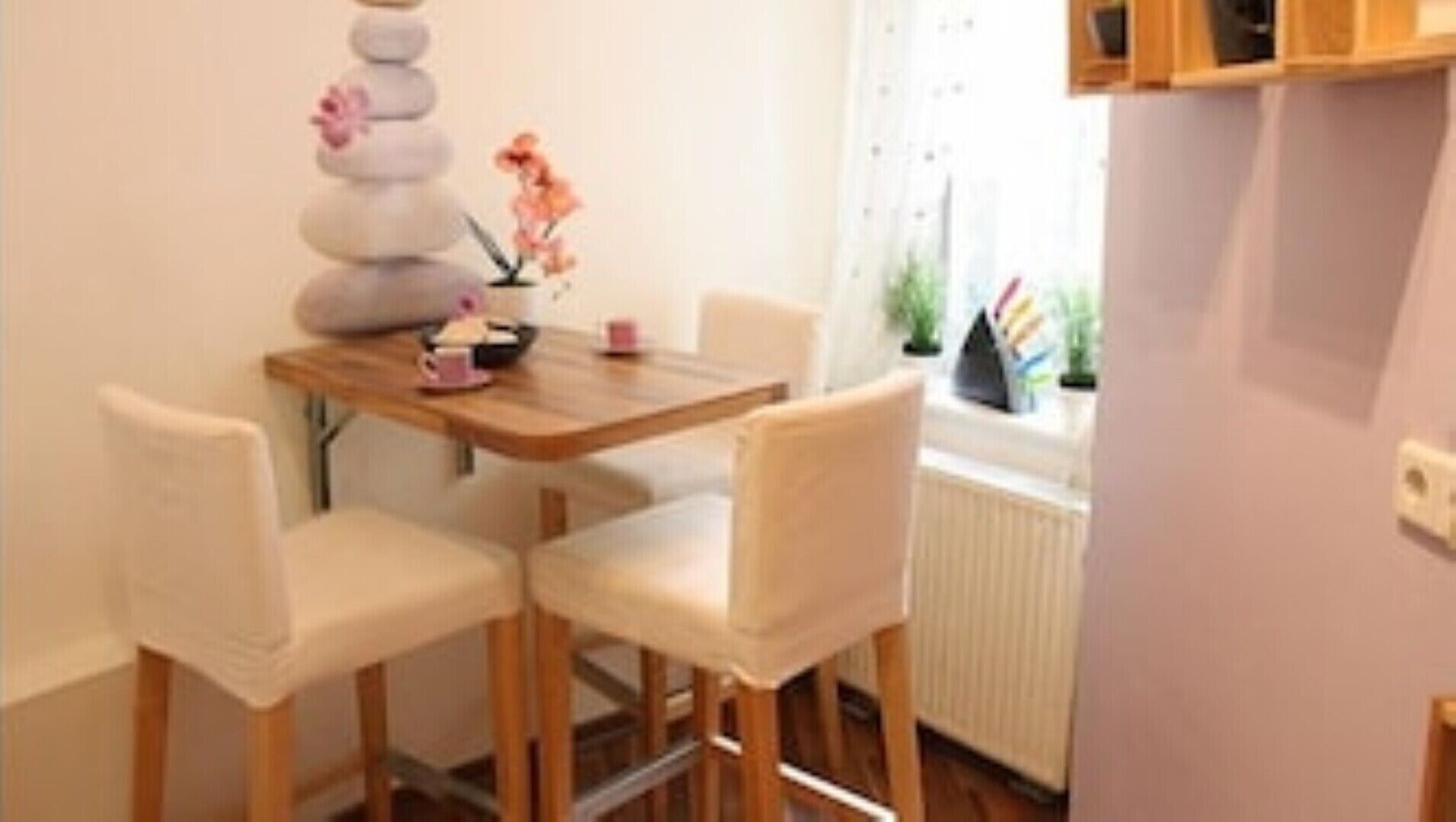 Un comedor con una mesa de madera y dos sillas blancas. Hay una pila de piedras con flores encima, un jarrón pequeño rosa y un objeto negro sobre la mesa.