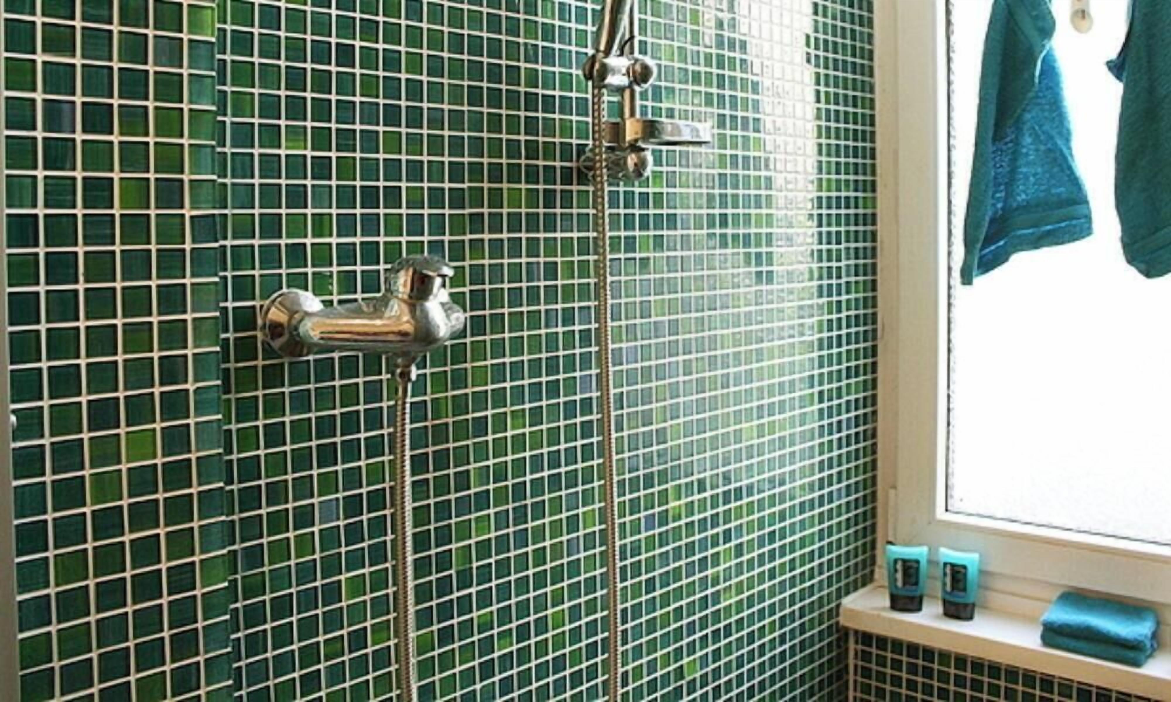 Un baño con mosaicos verdes, un cabezal de ducha, un perchero con dos toallas y una ventana con vistas.
