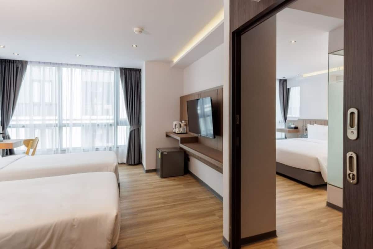 A modern hotel room with two beds, a TV, a small desk with a chair, and a view of the city through large windows.