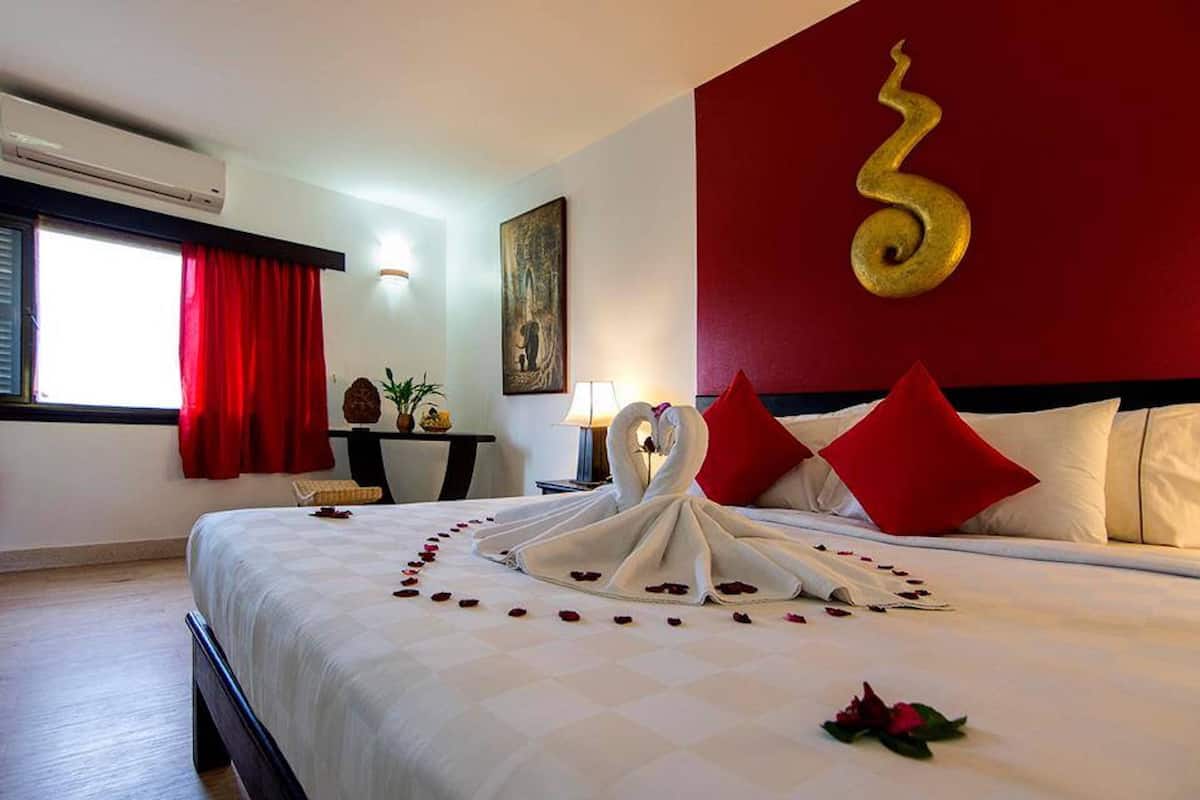 Habitación de hotel con cama, cortinas rojas, aire acondicionado instalado en la pared, un cuadro, una lámpara y un adorno de pared.