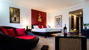 A hotel room with a bed, a wicker sofa, a small table with a bottle, and a wall with a red accent and a golden spiral design. - Le Siddharta D'Angkor (Siem Reap)