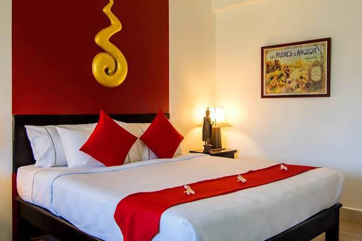 Una habitación de hotel con una cama, almohadas rojas, una colcha roja, una pared con diseño rojo y dorado, una mesita de noche con una lámpara y un cuadro enmarcado en la pared.