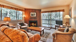 A cozy living room with a fireplace, comfortable seating, and large windows offering a view of trees. - Snow Flower 148 2 Bedroom Condo by RedAwning (Park City)