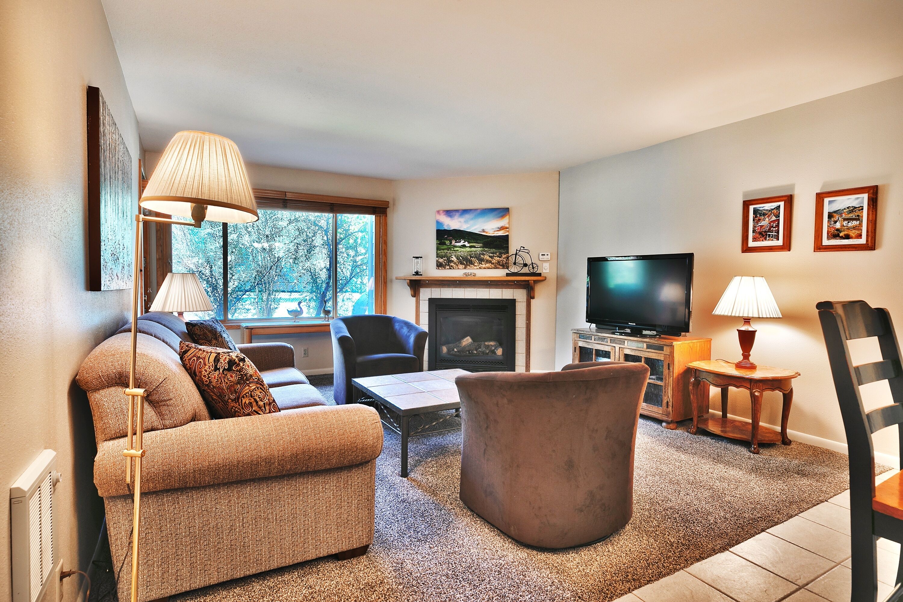 A living room with a sofa, armchair, ottoman, fireplace, television, and a window with a view of trees.