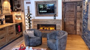 A cozy living room with a fireplace, two grey armchairs, a TV mounted above it, and wooden cabinetry. - Direct access to the slopes & village center; parking included. South-facing terrace. Cozy & comfortable 3* (Courchevel)