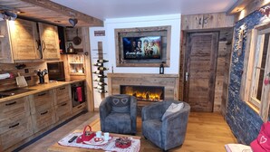 A cozy living room with a fireplace, comfortable seating, and a kitchen area with wooden cabinets and a wine rack. - Direct access to the slopes & village center; parking included. South-facing terrace. Cozy & comfortable 3* (Courchevel)