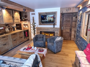 Un salón acogedor con chimenea, asientos cómodos y una zona de cocina con armarios de madera y una estantería para vinos.