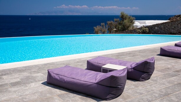 A poolside area with a view of the sea, stone flooring, and purple cushioned seating. - Villa Aliki  Panoramic view  Mykonos (Mykonos)