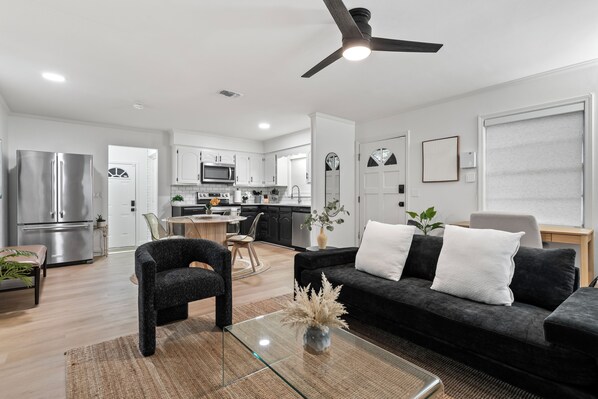A modern living room with a glass coffee table, a black sofa with white cushions, a dining area with a wooden table, and a kitchen with white cabinets and stainless steel appliances.