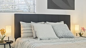 Een net opgemaakt bed met beddengoed met een patroon en meerdere kussens, een nachtkastje met een lamp en een klein plantje, en een raam met jaloezieën.