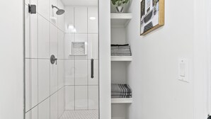 Een moderne badkamer met witte tegels, een douche met handdouche, toilet en wastafel. Er is een plank met opgevouwen handdoeken en een ingelijste foto aan de muur.