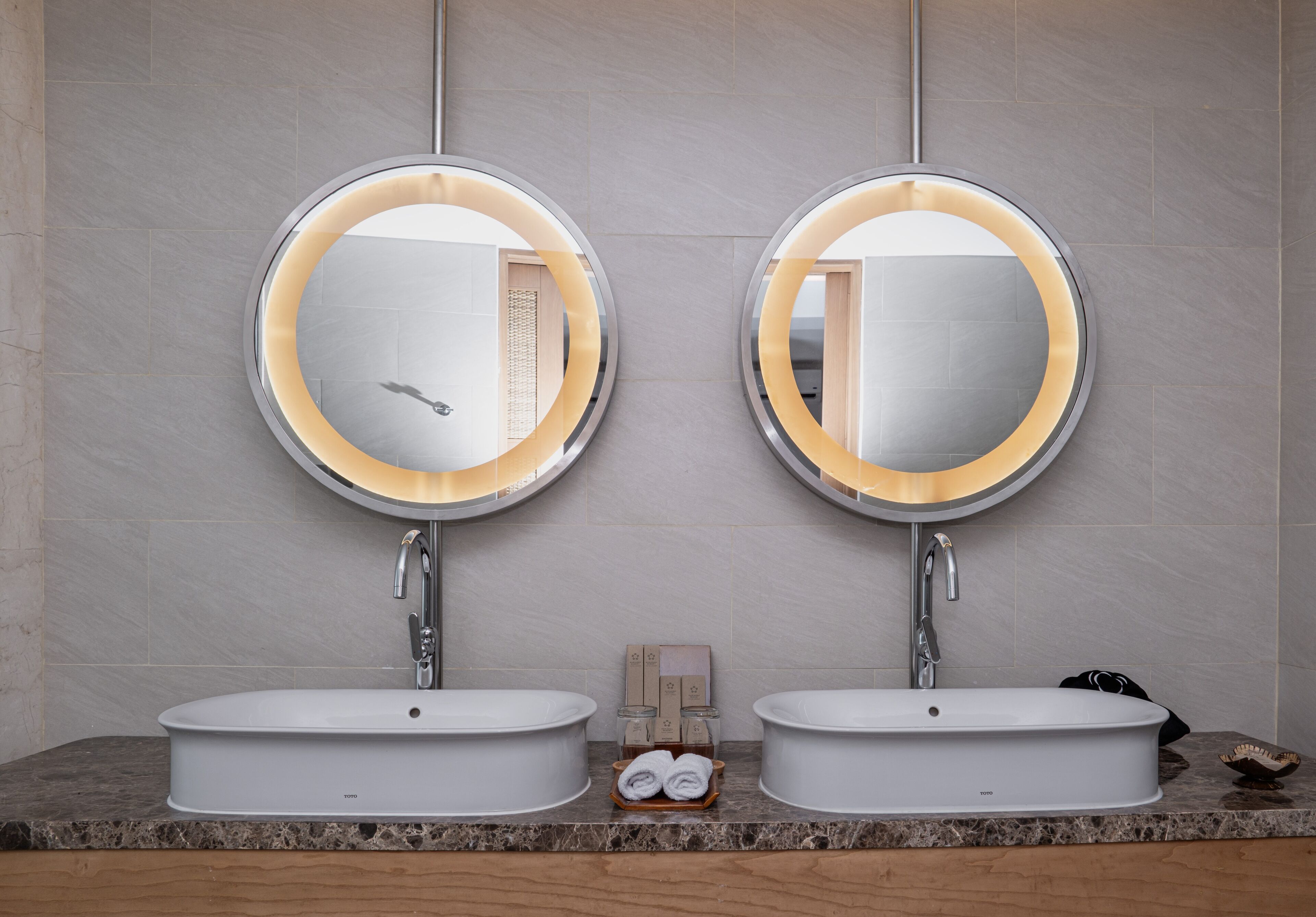 A bathroom with two sinks, each with a round mirror and a faucet. There is a wooden tray with towels and a soap dispenser between the sinks.