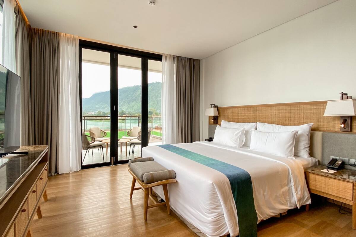 A hotel room with a large bed, a desk with a flat-screen TV, a balcony with a view of mountains, and a wooden floor.