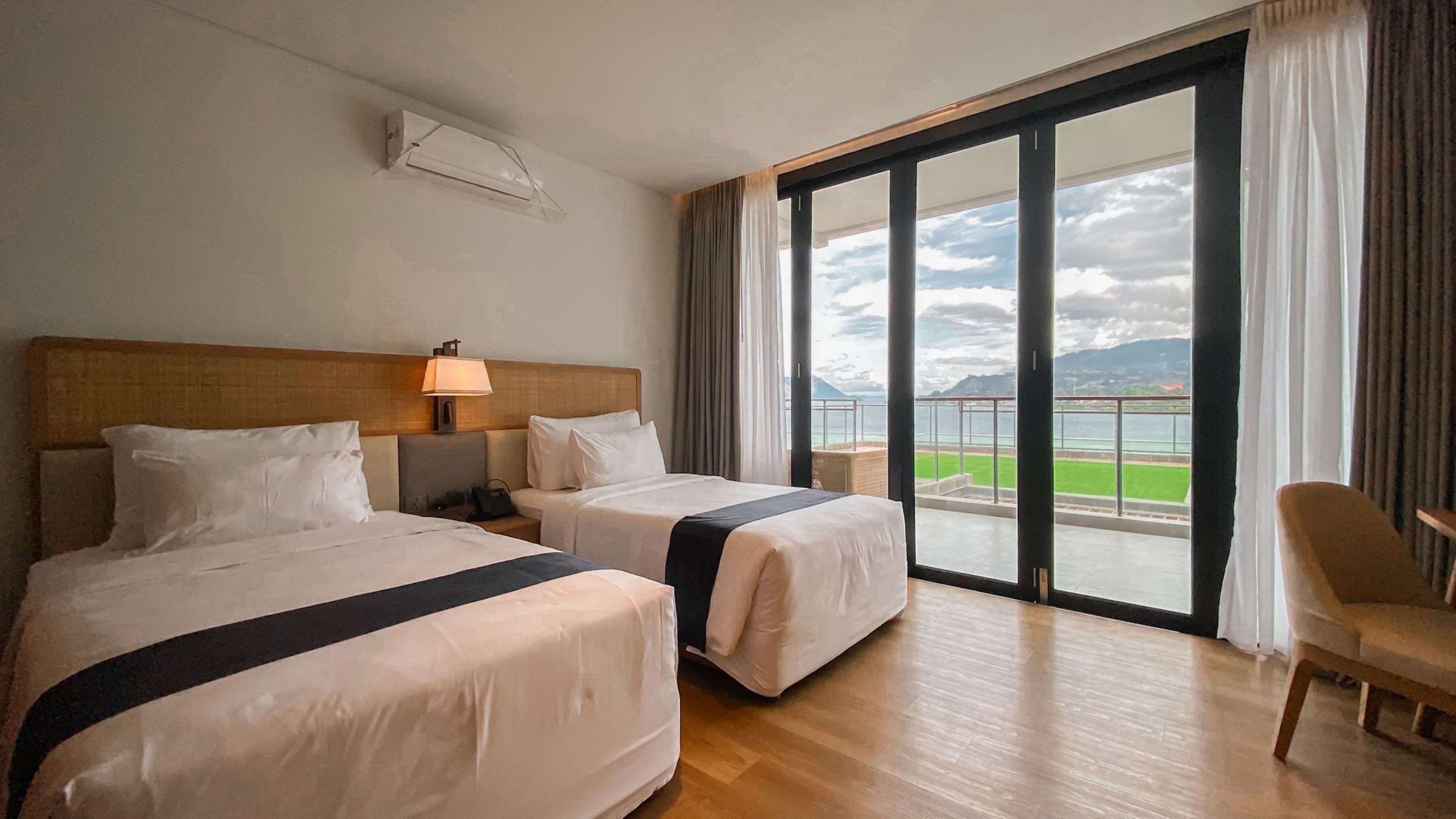 A hotel room with two beds, a nightstand, a lamp, and a large window offering a view of a sports field and mountains.
