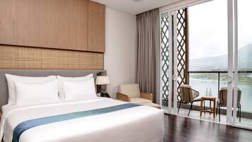 Habitación de hotel con una cama grande, cabecera de madera, balcón con sillas y vista a un lago.