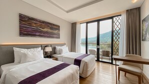 Camera d'albergo con due letti, una scrivania e una sedia. Ampia finestra con vista su una montagna e un lago.
