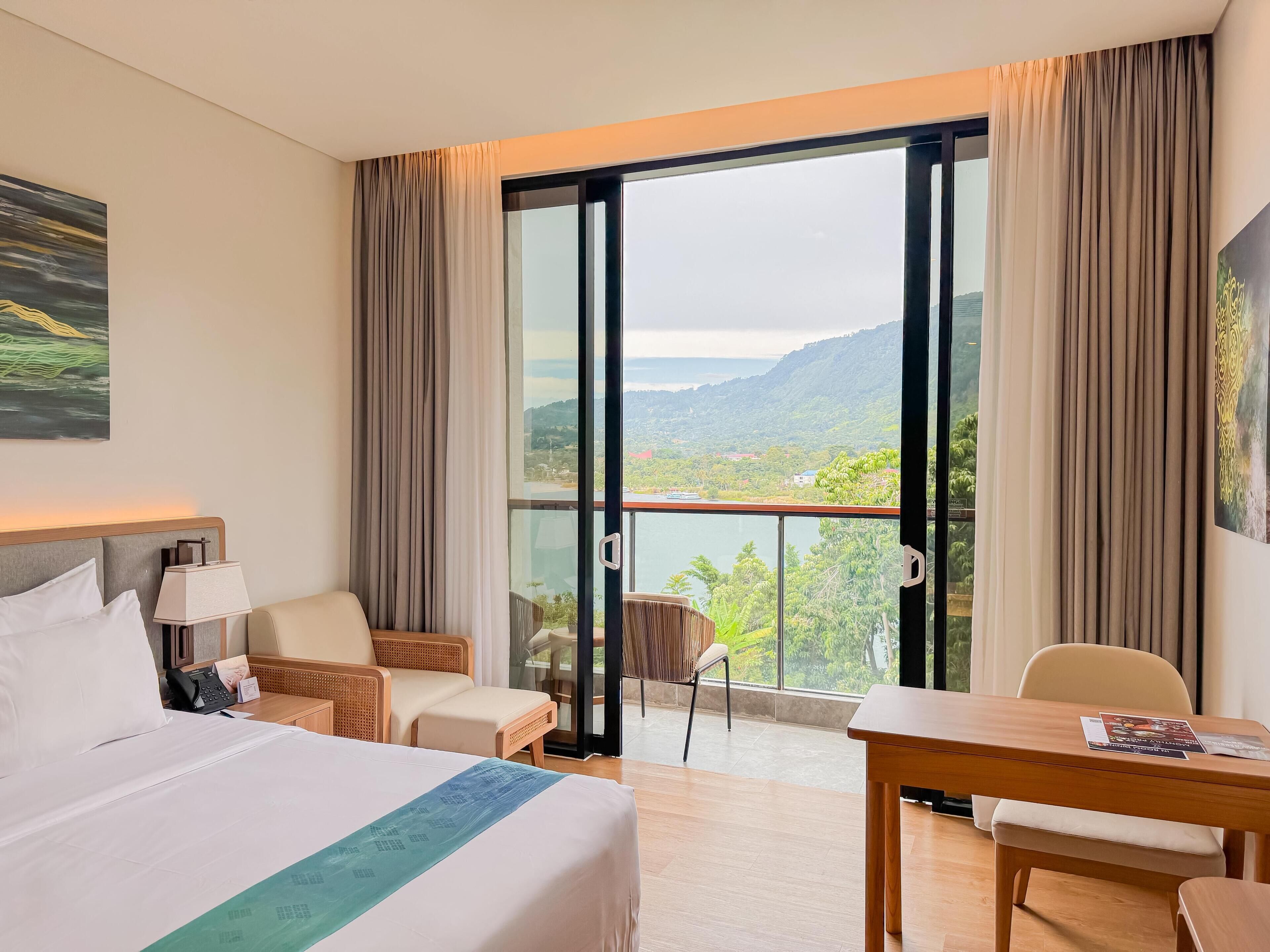 A hotel room with a bed, a desk, a chair, and a view of a lake and mountains.