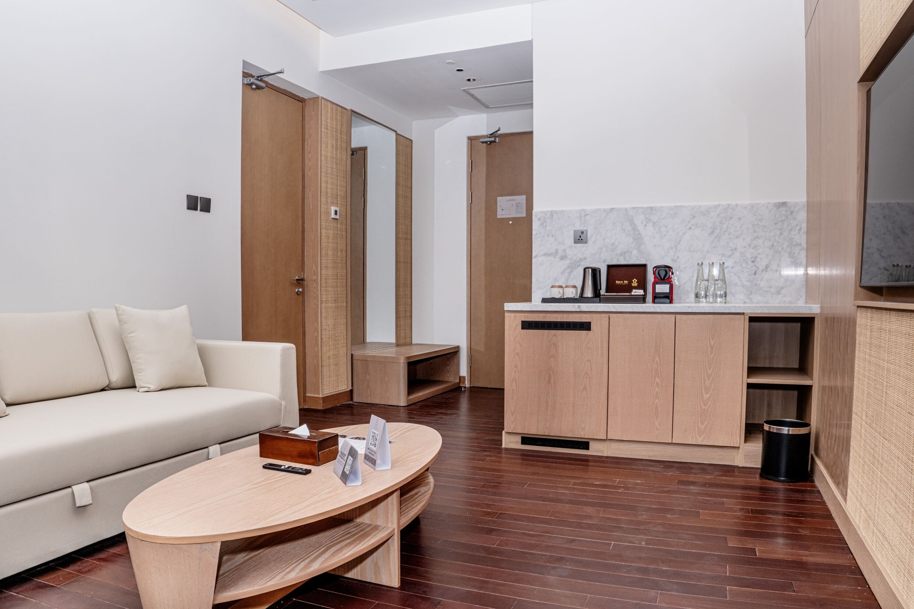 A modern hotel room with a wooden floor, a beige sofa, a wooden coffee table, a kitchen area with marble backsplash, and a TV mounted on the wall.
