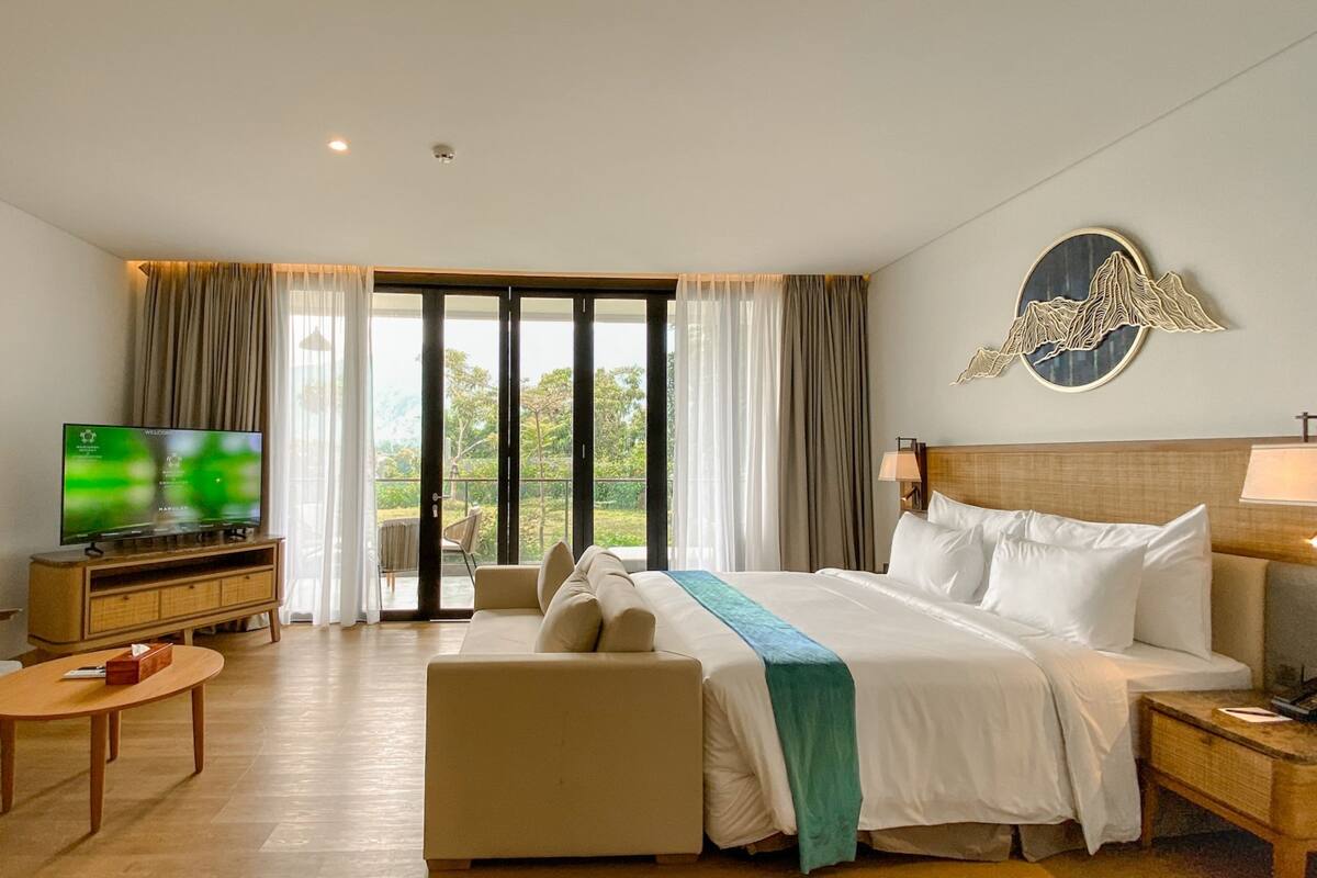 A hotel room with a bed, a TV, a sofa, a coffee table, and a view of greenery through large windows.