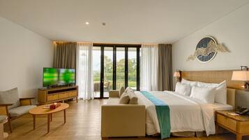 Habitación de hotel con cama, televisor, sofá, mesa de centro y vistas a zonas verdes a través de amplios ventanales.