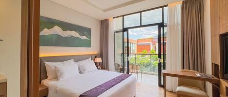 A hotel room with a large bed, a desk, a chair, a TV, and a balcony with a view of greenery and buildings.