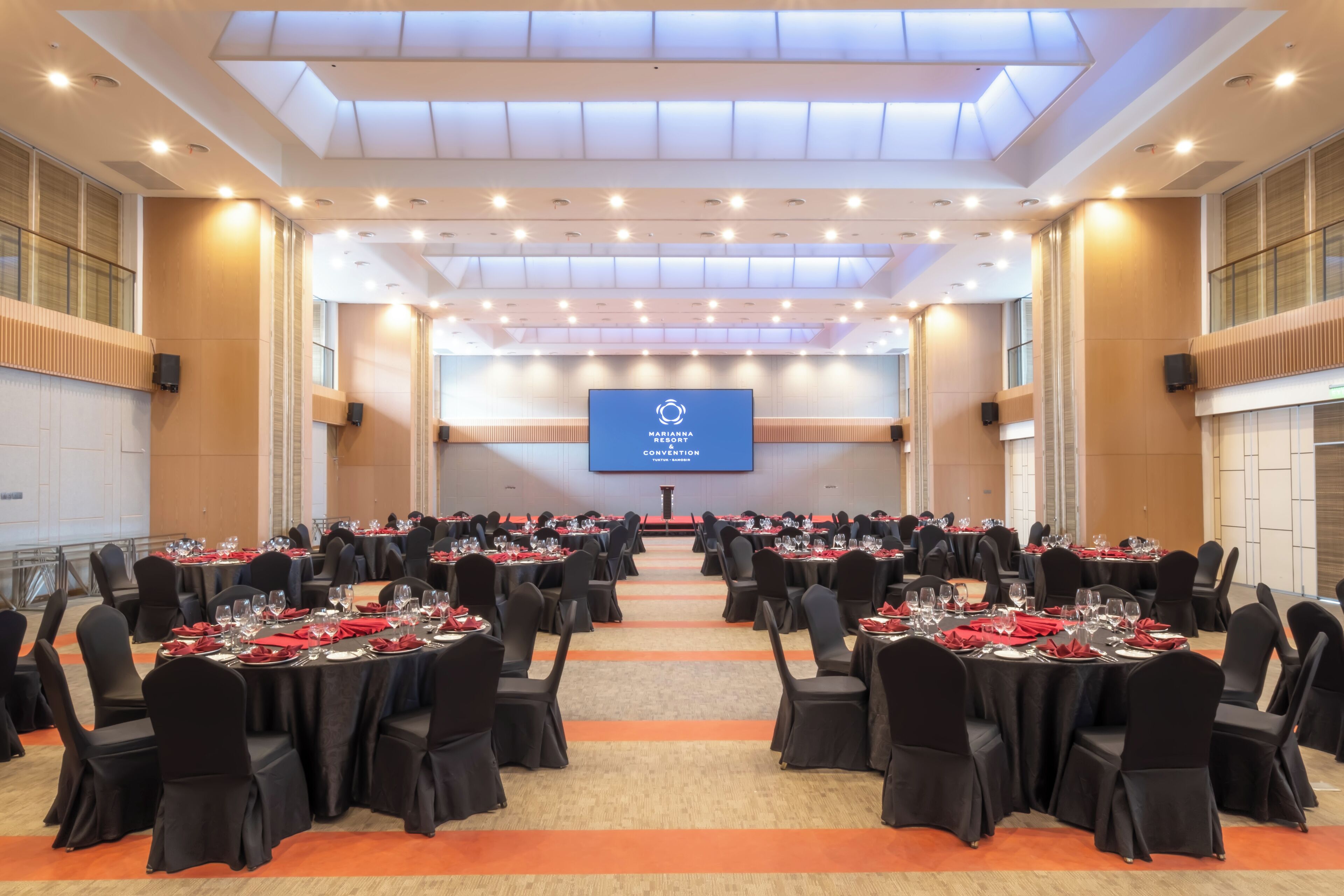 A large conference room with rows of tables set for a formal event, a stage with a screen, and a high ceiling with recessed lighting.