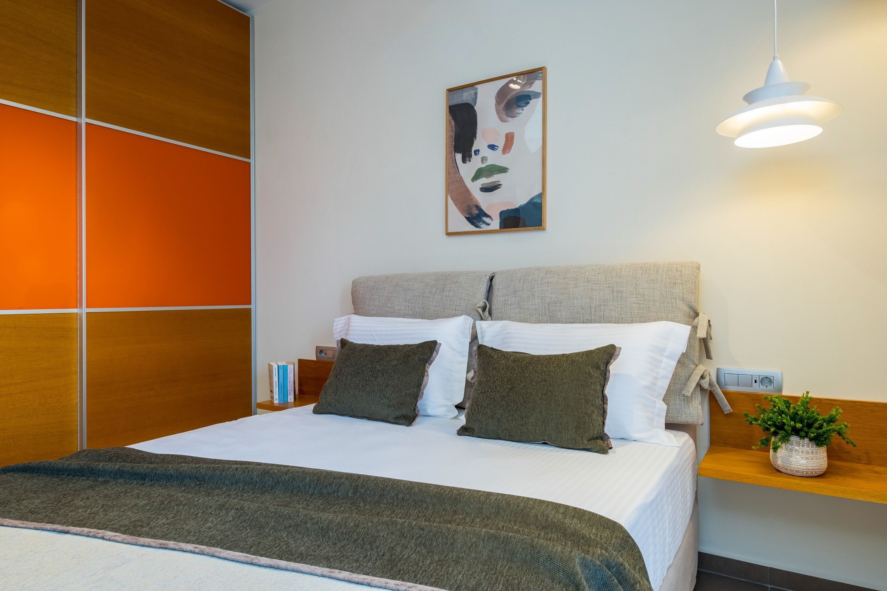 A modern hotel room with a large bed, a headboard with a wooden panel design, a bedside table with a plant, and a wall-mounted artwork.