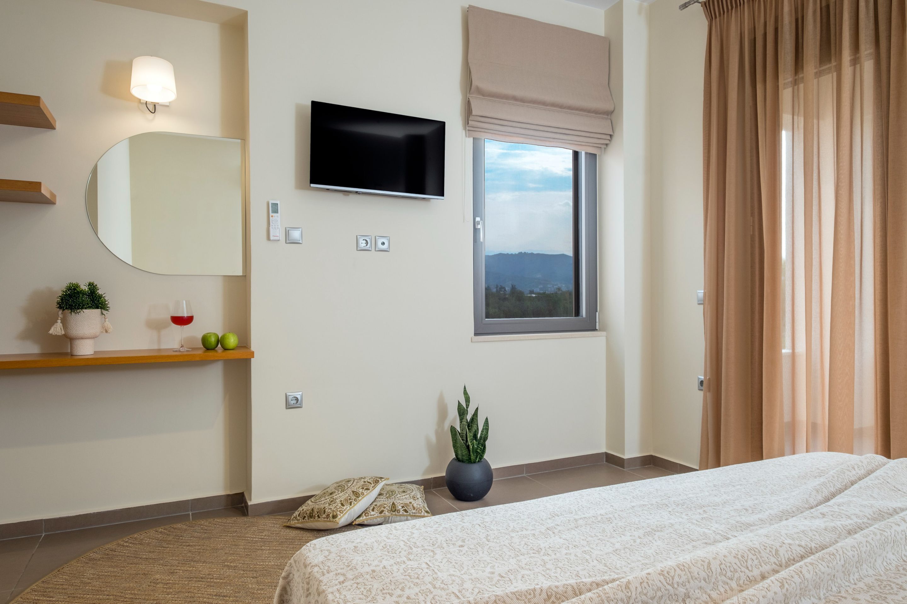 A bedroom with a bed, a TV, a window with a view, a mirror, and a shelf with decorative items.