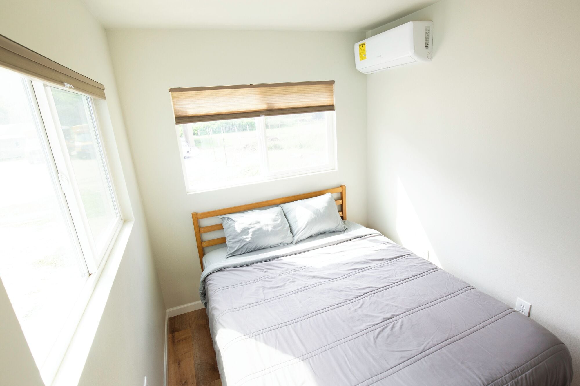 Una habitación pequeña con cama individual, cabecera de madera, ventana con persianas y equipo de aire acondicionado en la pared.