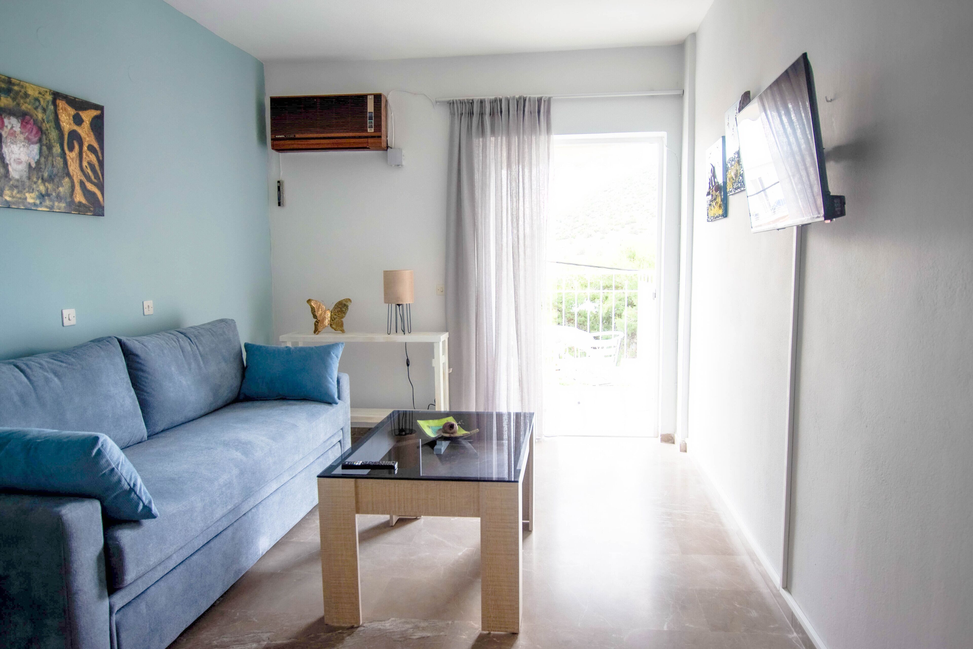 Una sala de estar con un sofá azul, una mesa de centro de vidrio y un televisor montado en la pared.