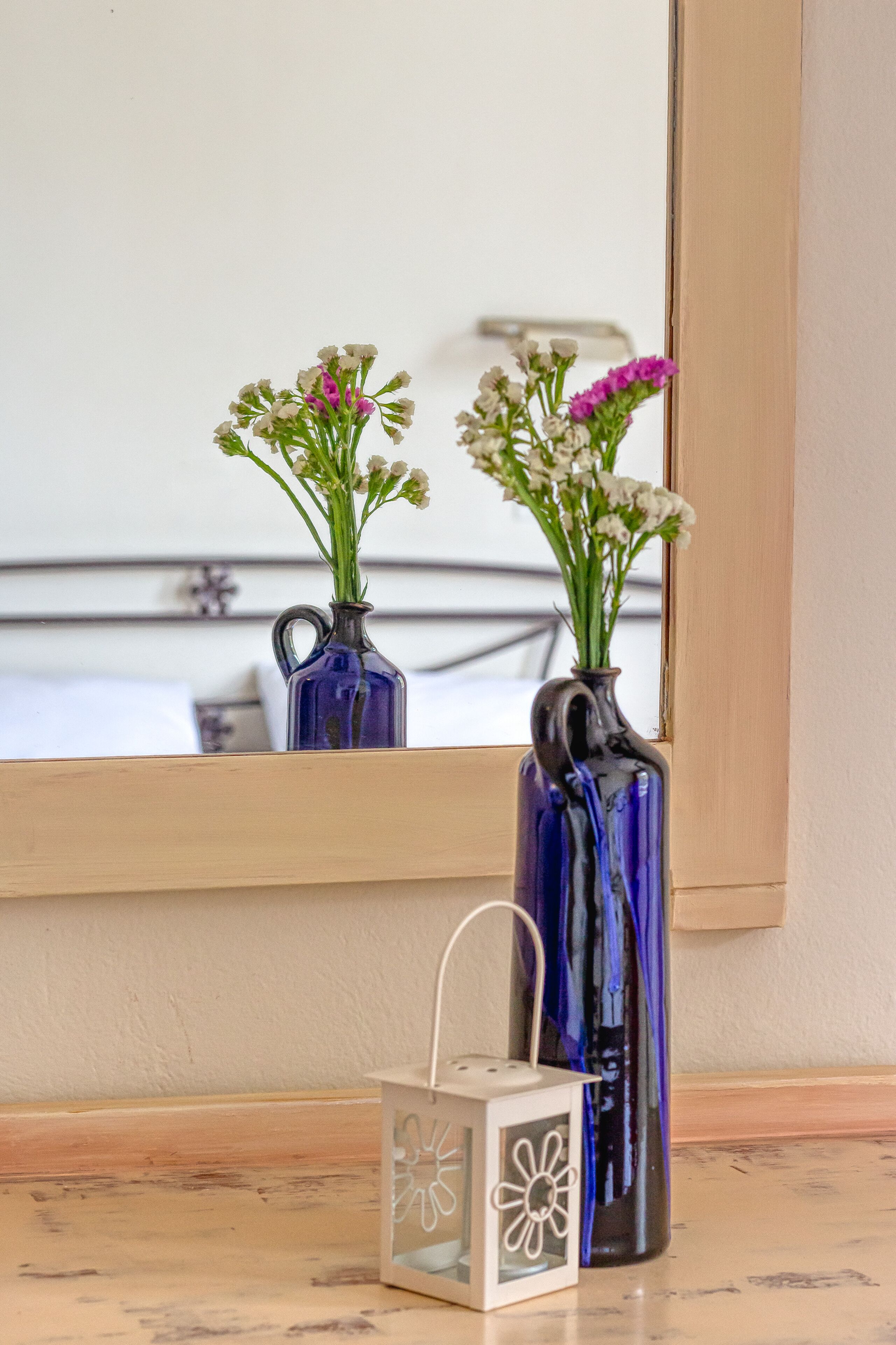 Twee vazen met bloemen op een vensterbank, een kleine lantaarn en een spiegel die een bed en raam weerspiegelt.