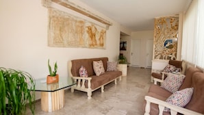 A living room with a brown sofa, a glass coffee table, and a white armchair. There is a large painting on the wall and a decorative panel on the right. - Akrotopi Apartments (Nafplio)
