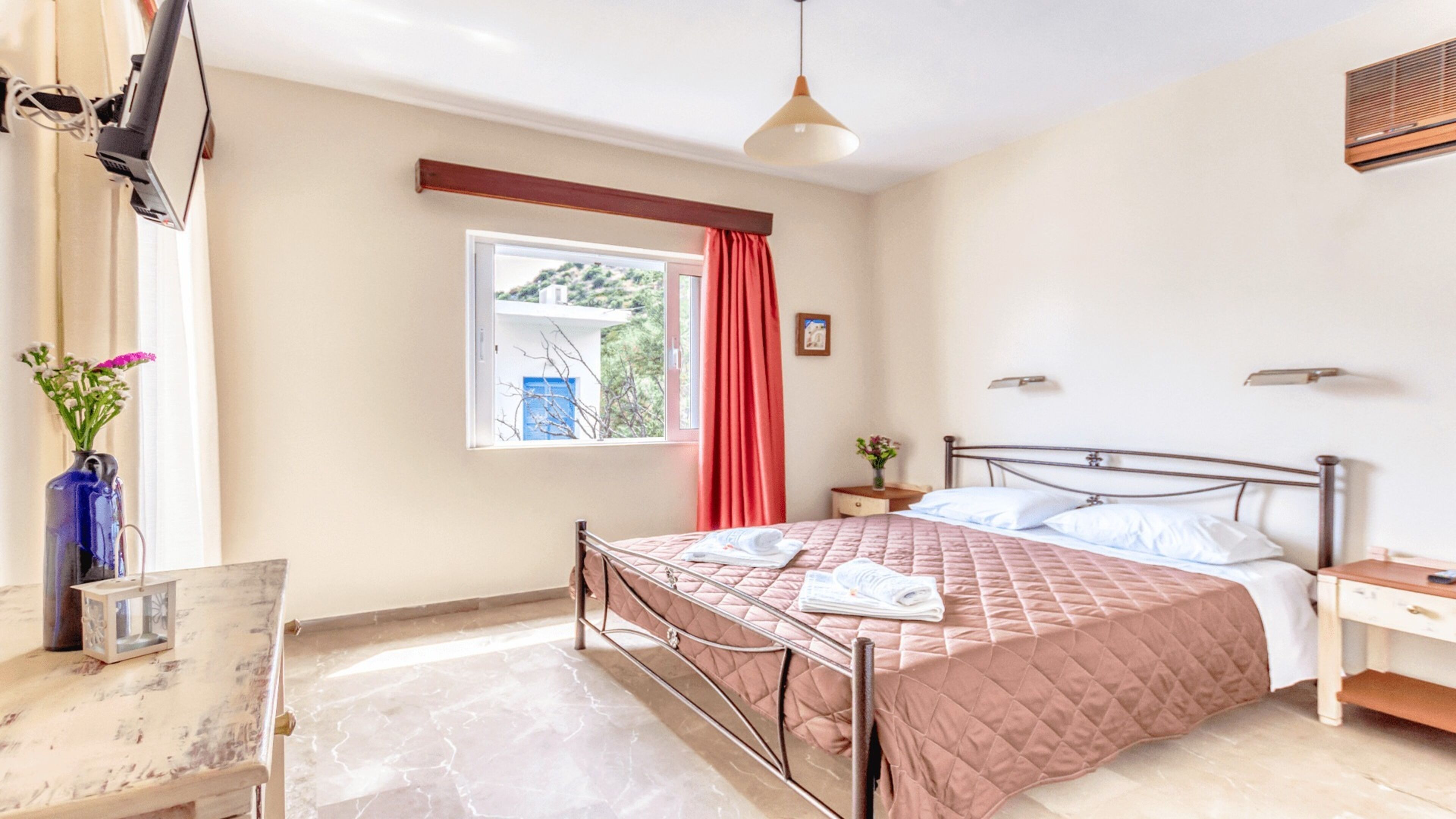 A bedroom with a bed, bedside tables, a desk, a window with curtains, and a hanging light fixture.