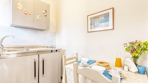 A kitchen with a sink, stove, and cabinets, a dining table with a chair, and a framed picture on the wall. - Akrotopi Apartments (Nafplio)