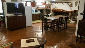 Amplia sala de estar con piso de madera en patrón de espiga, una mesa de comedor con sillas, un televisor, un refrigerador y una zona de cocina con microondas y horno.