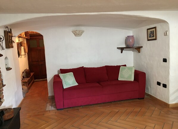 A red sofa with two pillows, a wooden floor, a white wall, a wooden door, and a shelf with a vase. - Traditional Sardinian semi-basement tavern - 5 min from the sea in Ogliastra (Girasole)