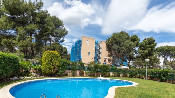 Una piscina con dos escaleras, rodeada de césped y árboles, y un edificio al fondo.