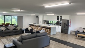 Een moderne woonkamer met een grijze bank, een salontafel en een eethoek met barstoelen. Op de achtergrond is een keuken met witte kasten en een roestvrijstalen koelkast zichtbaar.