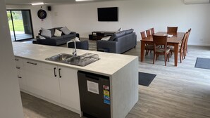 Een moderne keuken met een ingebouwde vaatwasser, een eethoek met een houten tafel en stoelen, een grijze bank en een flatscreen televisie aan de muur.