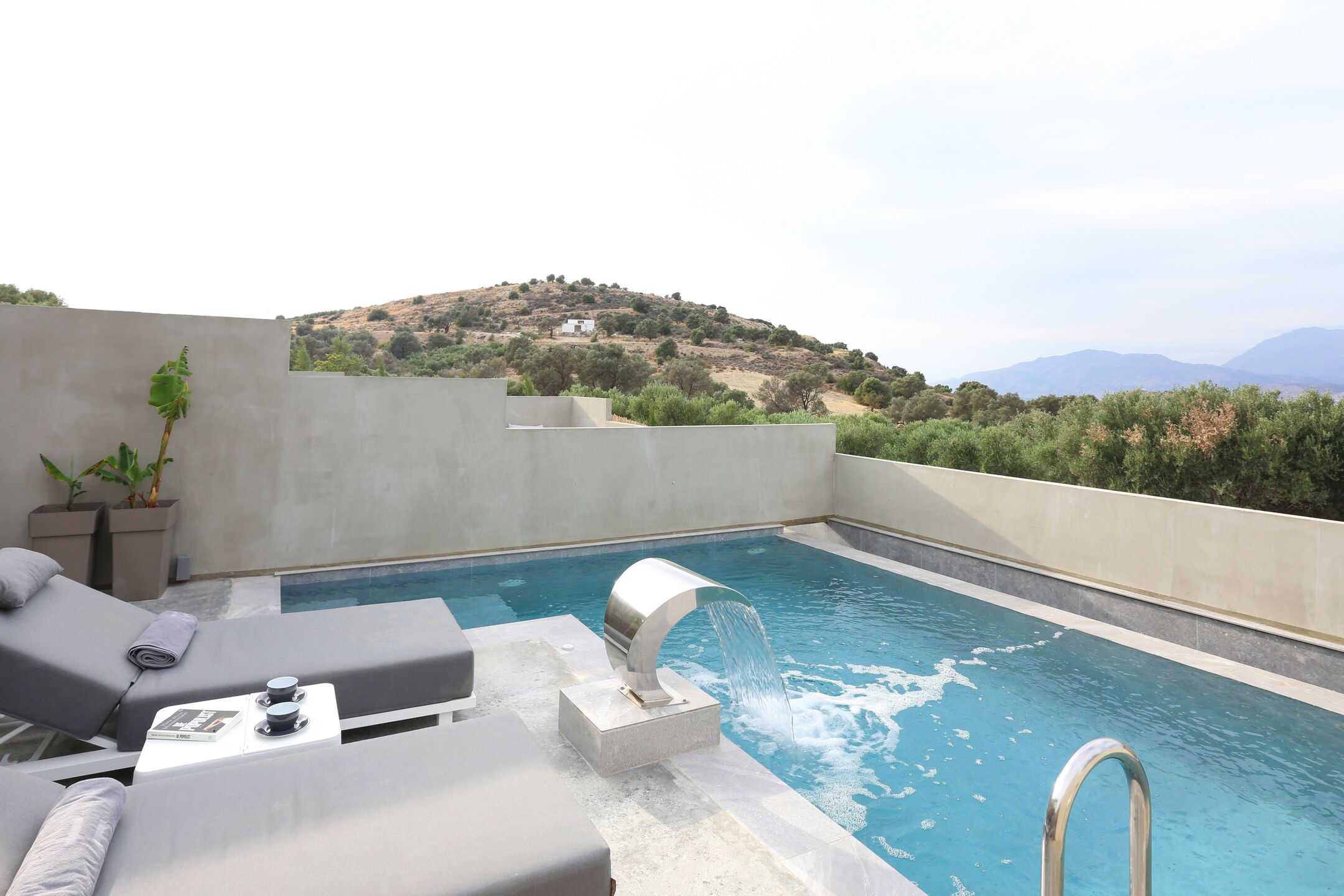 A rooftop pool with a waterfall feature, lounge chairs, and a view of a hilly landscape.