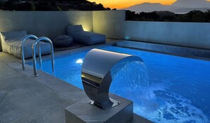 A modern outdoor pool with a water feature, seating, and a view of the mountains.
