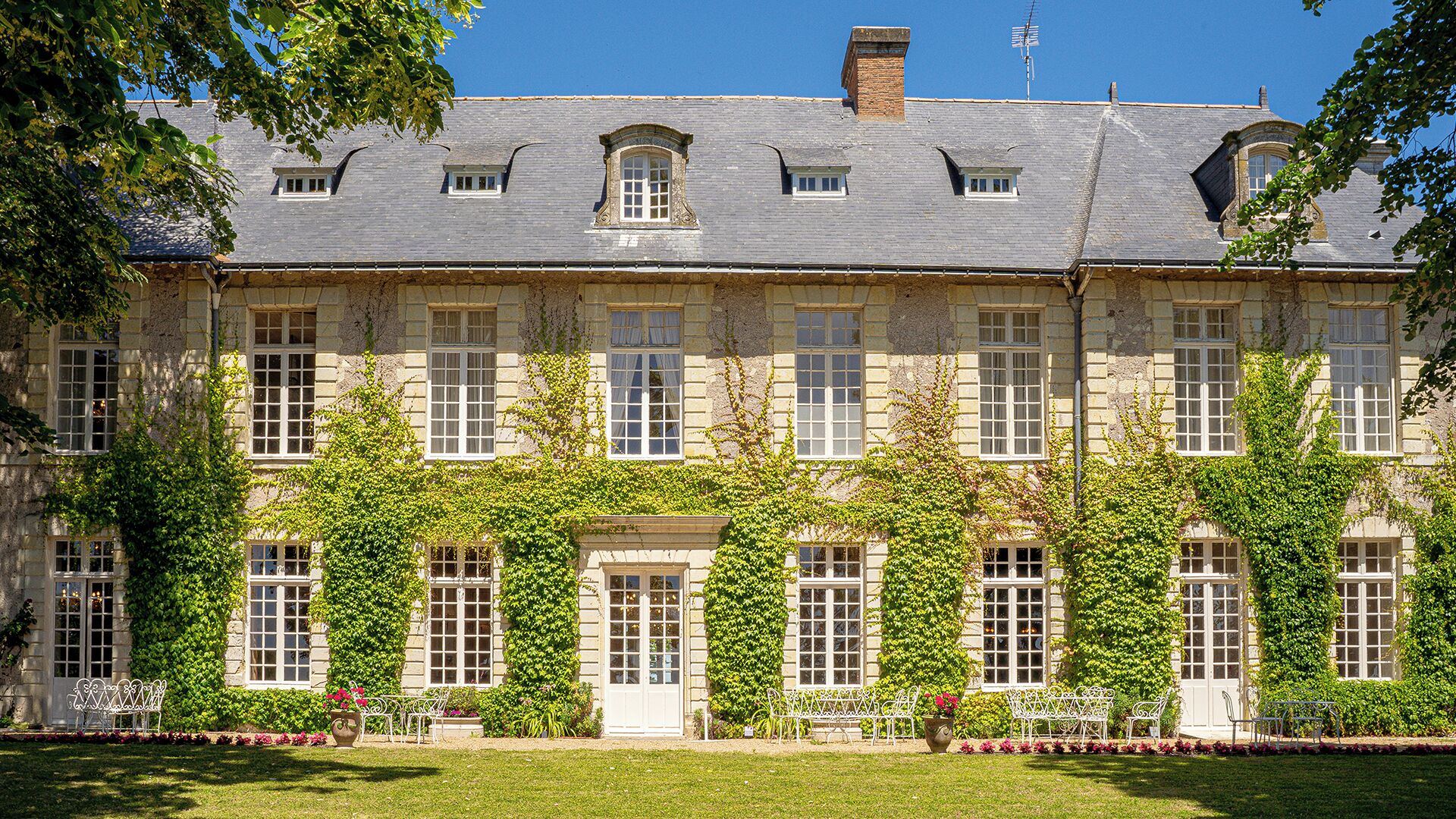 A large stone building with multiple windows and a symmetrical facade, surrounded by lush greenery and a well-maintained lawn.
