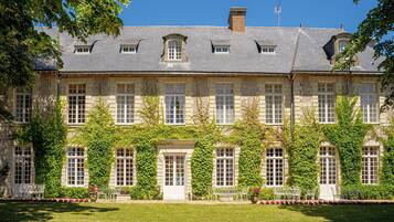 A large stone building with multiple windows and a symmetrical facade, surrounded by lush greenery and a well-maintained lawn.