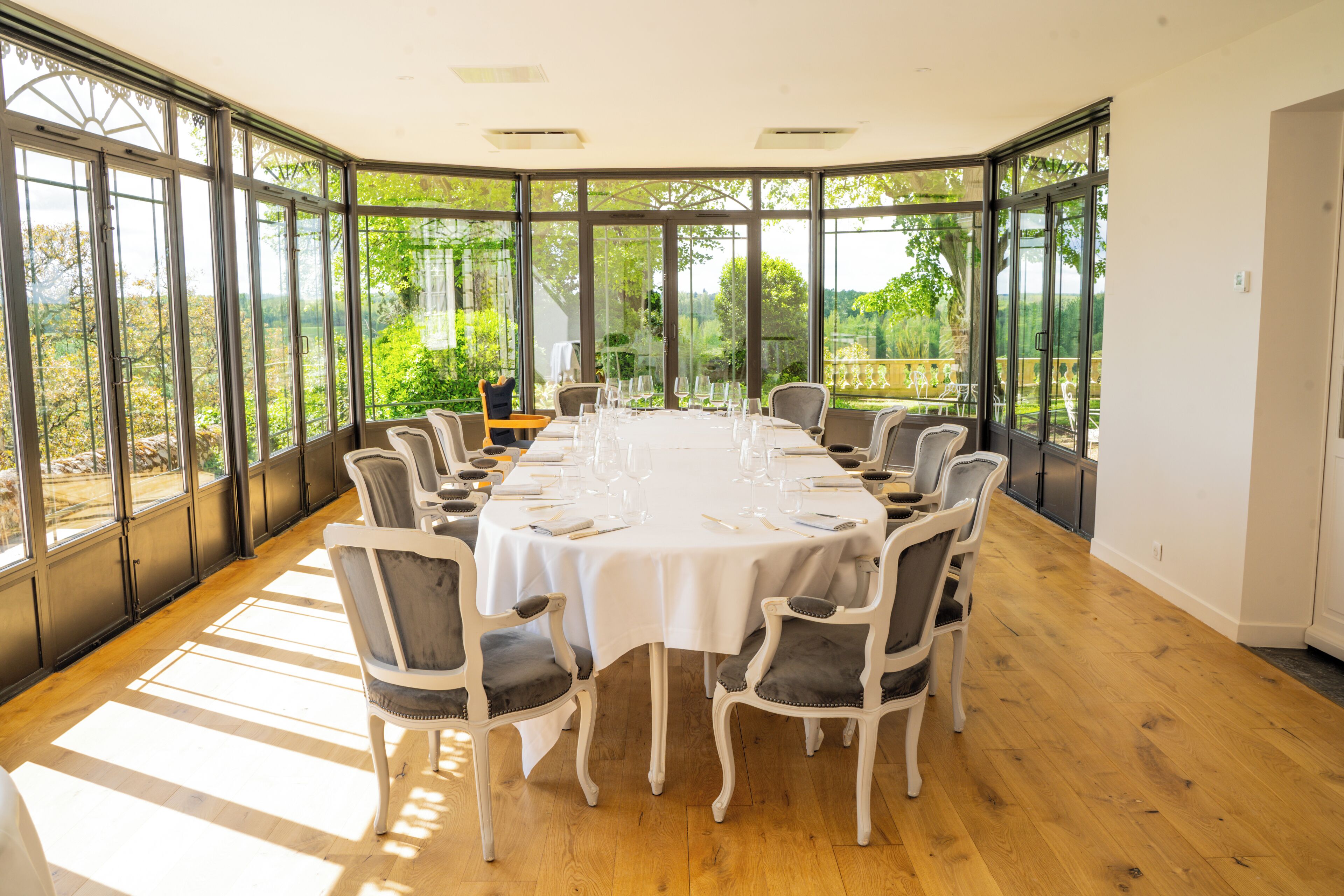 A large, round dining table set for six with white tablecloths and grey upholstered chairs, surrounded by large windows offering a view of greenery.